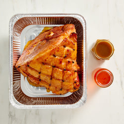 Denningers Roasted ham in a metal tray with two small jars of glaze on a marble surface