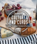 A styled cheese and wine board with grapes, a glass of red wine, a croissant, blue cheese, brie, and a bottle on a white fabric background. A large white oval logo overlay reads 'Denninger's Culture and Curds: A Guide to Perfect Pairings & Platings'.