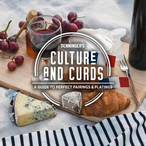 A styled cheese and wine board with grapes, a glass of red wine, a croissant, blue cheese, brie, and a bottle on a white fabric background. A large white oval logo overlay reads 'Denninger's Culture and Curds: A Guide to Perfect Pairings & Platings'.